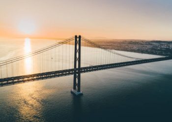 Aerial view suspension bridge over Tagus river in Lisbon. Drone flying above water opposite 25th april bridge symbol of Portugal in sun shining, beautiful landscape at sunset.